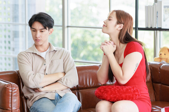 Asian young beautiful female girlfriend sitting cuddling smiling on cozy sofa in decorated living room showing little finger reconciling handsome upset sulking male boyfriend at home on valentine day