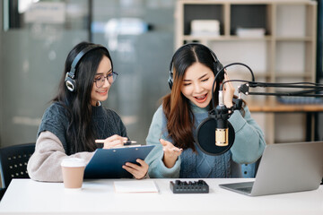 Young woman and Asian woman wearing headphones and doing a live podcast for their channel,...