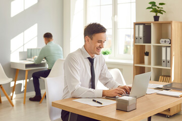 Portrait of cheerful happy smiling attractive business man wearing white shirt and tie typing on laptop sitting at the desk on workplace. Two male young company employees working in modern office. - obrazy, fototapety, plakaty