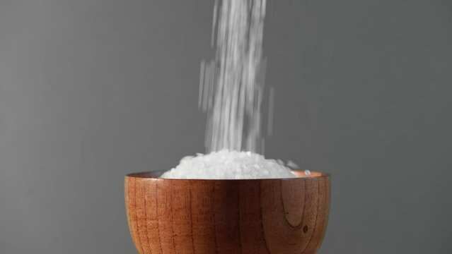 White table salt granules in a pile with spoon. Wood bowl. Gray color studio background. Condiment particles spinning. Clean cosmetic bath salt footage. Closeup of grained sea salt pieces hill	