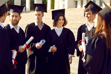 Group of a happy smiling graduate students in black graduation robes talking near university...