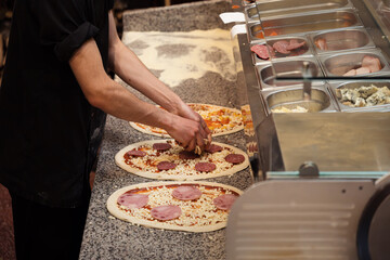 A chef in a black apron meticulously adds toppings to pizzas, with a stainless steel counter of...