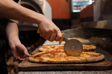 A chef's hand is shown slicing through a pizza with a rolling cutter on a wooden serving board
