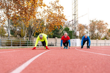 Group of three elderly male athletes poised at the start line on a running track, embodying...