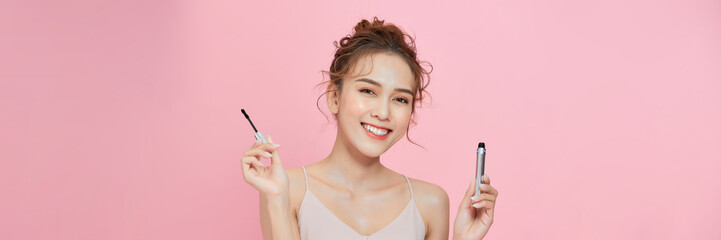 happy young woman holding mascara tube and brush isolated on pink background