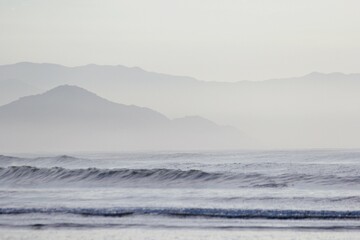 Blurred seascape with foggy morning on seashore. Light blue sea, small waves, mountains silhouette, cloudy sky.  