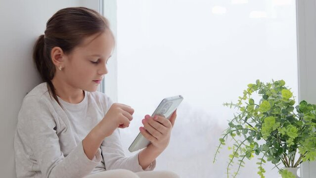 Adorable brown haired little girl relaxing on windowsill using mobile phone scrolling online looking at smartphone screen checking social network using gadget for leisure time at home