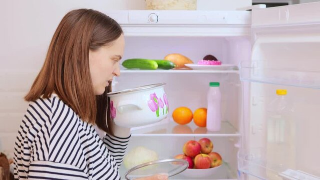 Brown haired woman wearing striped shirt opening fridge taking pot with spoiled soup frowning face grimacing from unpleasant aroma expressing negative emotions