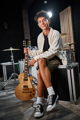 Fototapeta premium cheerful good looking teenage boy in casual attire holding guitar and smiling at camera in studio