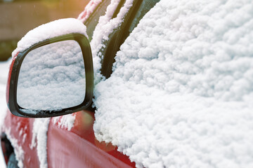 Fragment of red car covered with snow during snowfall. Mirror of car under snow. Car covered with...