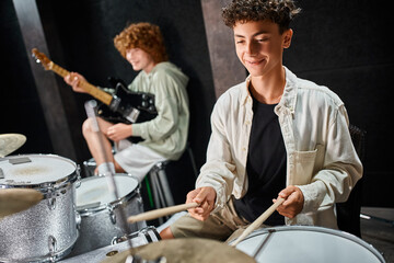 joyful adorable teenage boy with braces playing drums next to his friend with guitar, musical group