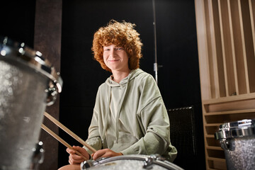 cheerful adorable teenager with red hair in casual outfit in front of his drum set smiling at camera