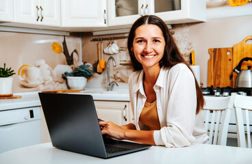 Pretty happy brunette woman using computer sitting at the kitchen table.