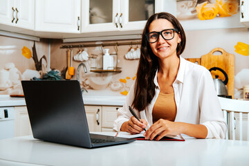 Portrait of happy female student watching webinar or tutorial studying online on laptop sitting at kitchen table, noting down after teacher or lecturer.