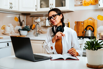 Portrait of happy female student watching webinar or tutorial studying online on laptop sitting at kitchen table, noting down after teacher or lecturer.