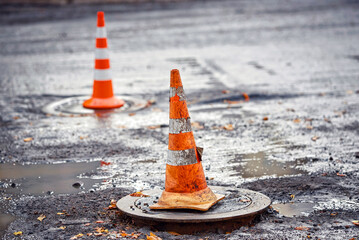 Orange traffic cone stands on manhole. Road repair works, asphalt laying, pylon to mark an obstacle or hole on road. Traffic cone stands on hatch, asphalt paving works - obrazy, fototapety, plakaty