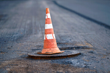 Orange traffic cone stands on manhole. Road repair works, asphalt paving, pylon to mark an obstacle or hole on road. Traffic cone stands on hatch. Road reconstruction, renew old asphalt surface - obrazy, fototapety, plakaty