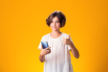 Surprised kid girl holding smartphone in hand showing thumbs up gesture over yellow background