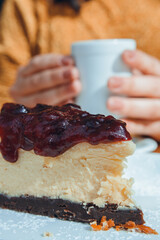 vertical image, slice of cheesecake with female hands holding cup of coffee blurred in background
