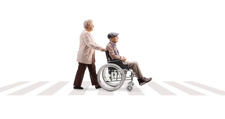 Full length profile shot of an elderly woman pushing an elderly man in a wheelchair at a pedestrian crossing - obrazy, fototapety, plakaty