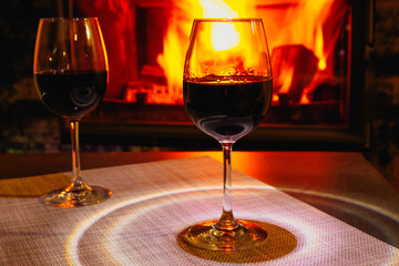 Two glasses of red wine against the background of a burning fireplace in hard reflective lighting. Romantic relaxed dinner by the fireplace.