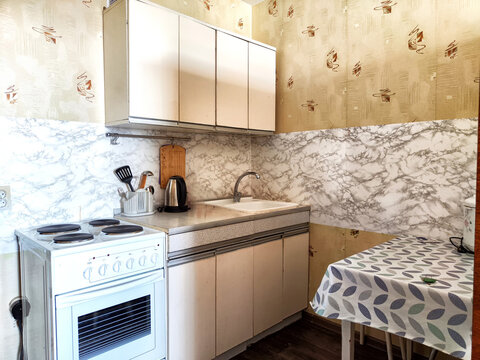 The interior is a small old kitchen with a table, stool, cabinets, sink and stove. Design in the Soviet poor outdated style