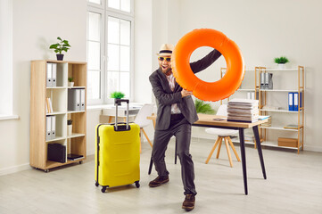 Young excited man in sunglasses with rubber ring and suitcase wearing office clothes having fun on...