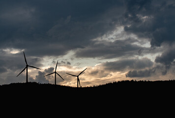Wind turbines on the hill, sunset with dark cloluds. Sky with silhouette of mountains and forest.
