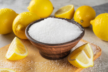 Citric or lemon acid in clay bowl with a spoon, close-up, and whole ripe lemons on gray background. 