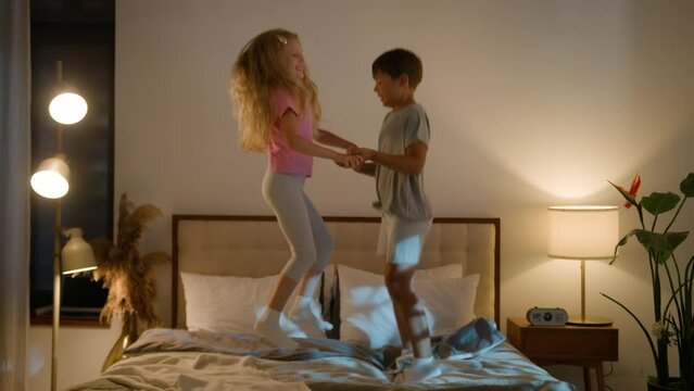 Active energetic laughing funny Caucasian children brother sister kids girl boy siblings jumping on bed play together at home evening having fun fooling around jump in bedroom bedtime happy childhood