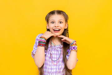 The child put on a lot of bracelets made of beads and beads. A happy little girl is enjoying a beaded ornament. Beading for children. Isolated yellow background. - obrazy, fototapety, plakaty