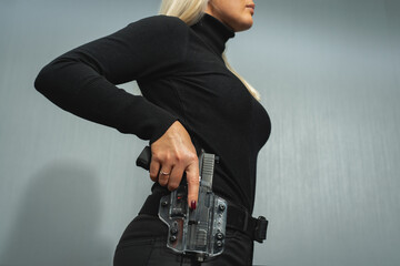 A girl in black clothes pulls out a pistol from a transparent holster on her belt, close-up photo.