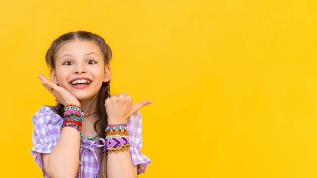 Naklejki A little girl points her finger at your advertisement. The child put on a lot of beaded bracelets. Beading for children. Yellow isolated background. Copy the space.