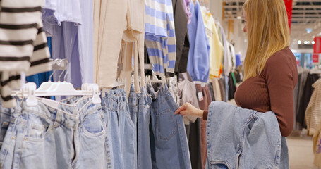 A young woman chooses jeans in a clothing store - obrazy, fototapety, plakaty