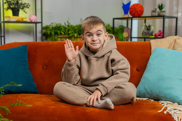Young child boy smiling friendly at camera and waving hands gesturing hello, hi, greeting or goodbye, welcoming with hospitable expression. Teenager Caucasian kid at home in living room sits on sofa