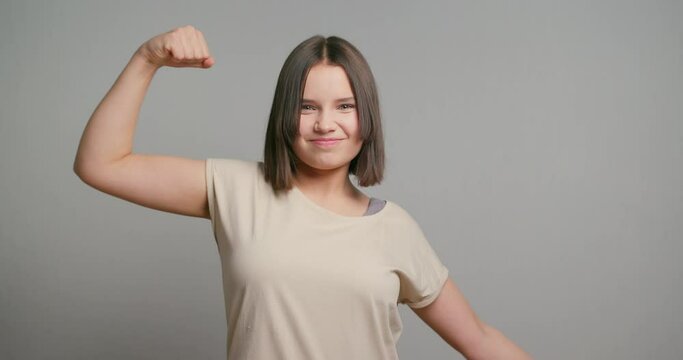Young strong latin teenager girl feeling happy, satisfied and powerful, flexing fit and muscular biceps, looking strong on gray background