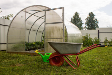 A garden cart and watering can on the background of a transparent plastic greenhouse on green...