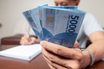 Close up of man's hands counting Indonesian rupiah notes and making notes, money financial...