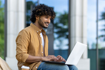 Close-up photo of a smiling young Indian man using a laptop while sitting on a street bench