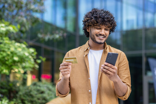 Close-up photo of a smiling young Nidhi man standing in the middle of the street using a mobile phone and credit card