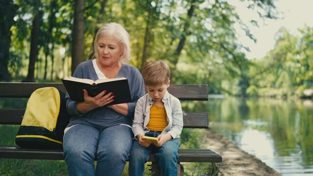 Senior lady reading book, little boy playing on gadget in park, generation gap