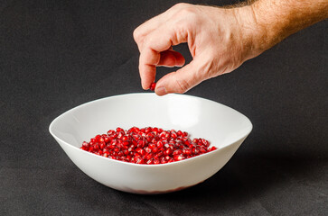 Man's hand throws pomegranate seeds into a white bowl with red pomegranate seeds. Black background.