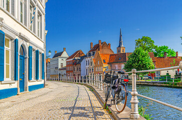 Bike near metal fence, embankment street of Sint Annarei water canal, buildings in Brugge old town, Bruges city historic center in sunny day, Saint Anna Church spire, West Flanders province, Belgium - obrazy, fototapety, plakaty