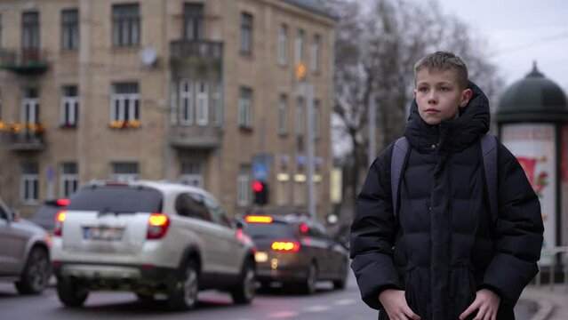 Schoolboy with backpack waiting for bus after lessons to go home. Winter season, boy wears warm jacket. Public transport stop.