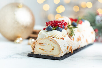 Meringue roll sprinkled with powdered sugar decorated with blueberries, pomegranate and almond flakes on the white background with blurred bokeh lights. Christmas festive pastry