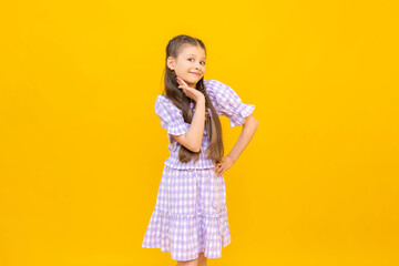 A charming little girl, adoringly clasping her hands in front of her face. The child is smiling blissfully on a yellow isolated background. A happy young girl.