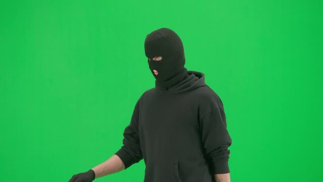 Portrait of thief on chroma key green screen background. Man robber wearing hoodie, jeans and black balaclava, walking getting ready for making a crime. Half turn.