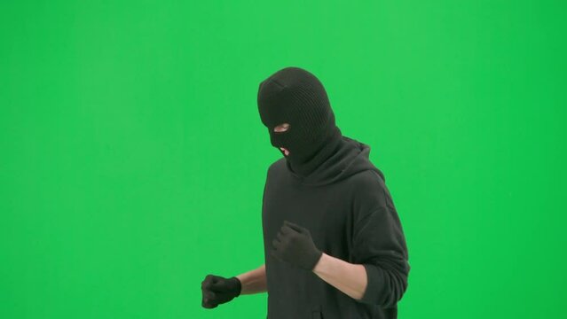 Portrait of thief on chroma key green screen background. Man robber wearing hoodie, jeans and black balaclava, running getting ready for making a crime. Half turn.