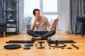 A man sitting at home on the floor is engaged in self-assembly of furniture from a set of parts in...