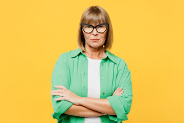 Elderly sad frowning blonde woman 50s years old wears green shirt glasses casual clothes hold hands crossed folded look camera isolated on plain yellow background studio portrait. Lifestyle concept. - obrazy, fototapety, plakaty